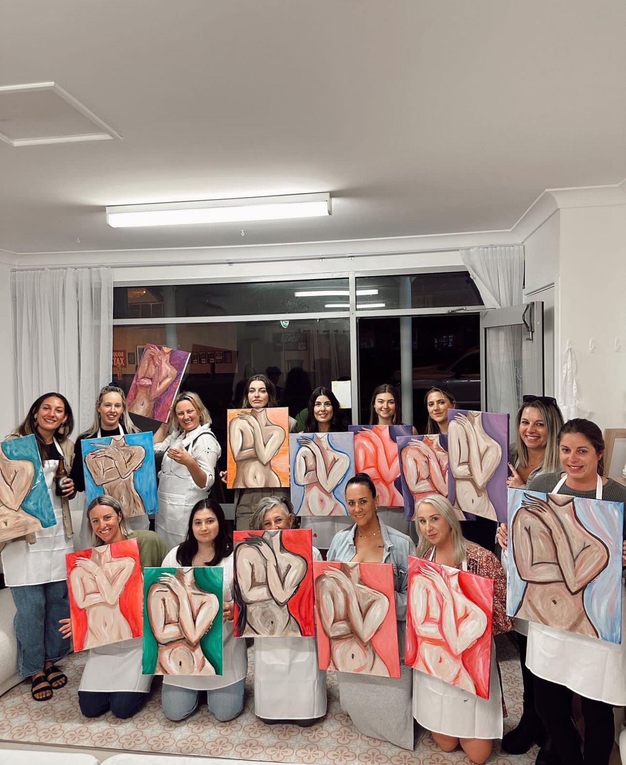 A group painting class shows off their completed figure artwork while wearing aprons.