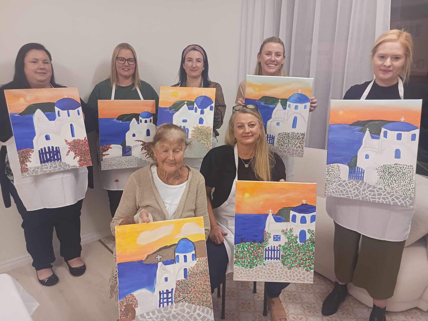 Art class participants display their completed paintings of a Mediterranean seaside church with blue domes.