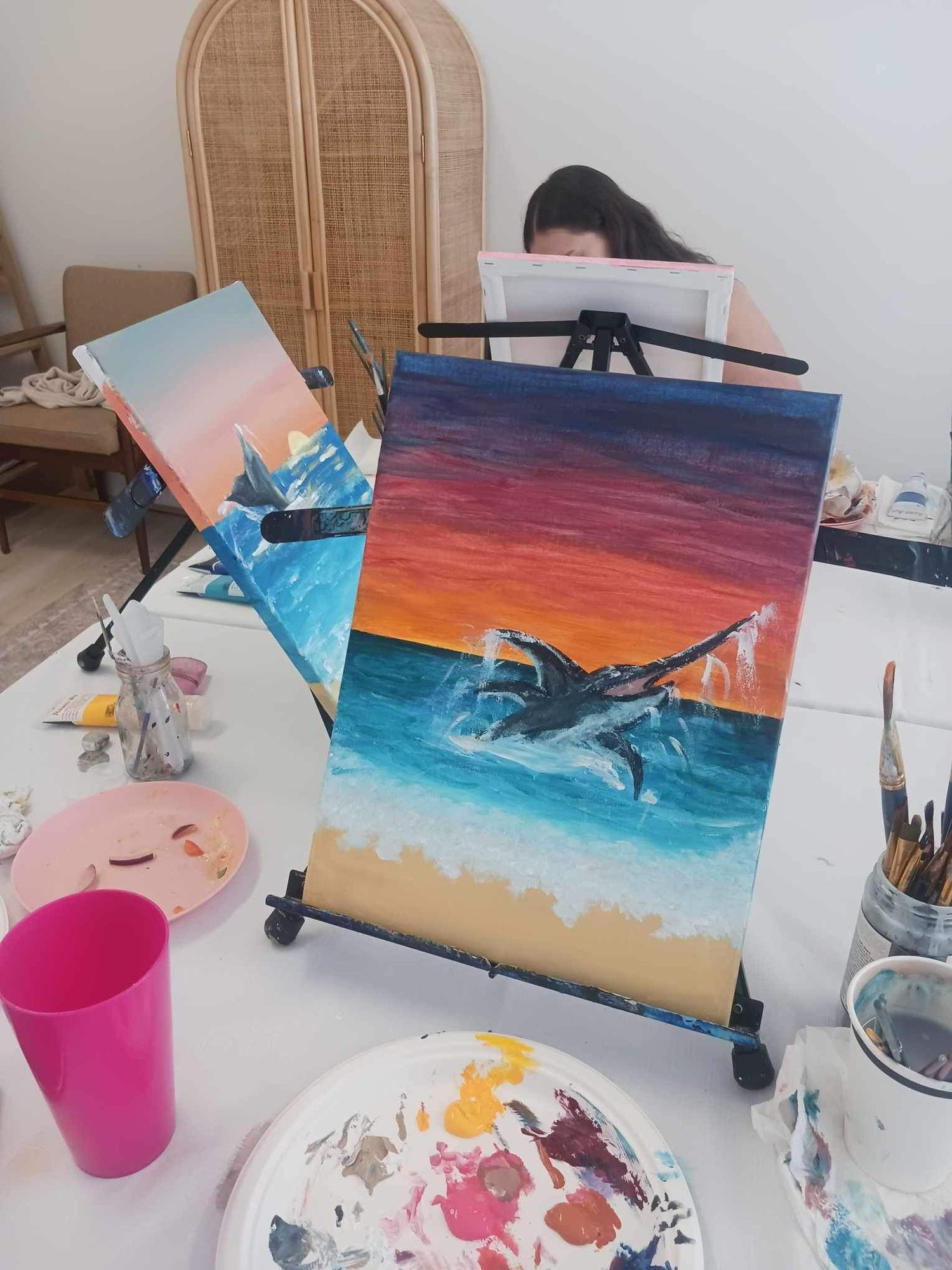 Colorful ocean sunset painting with jumping dolphin on easel surrounded by art supplies.