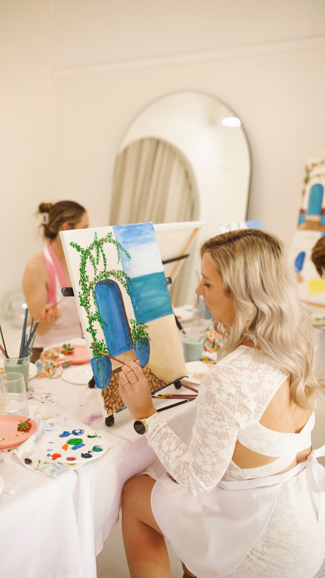 Woman in white lace examining her beach-themed artwork during a painting class.