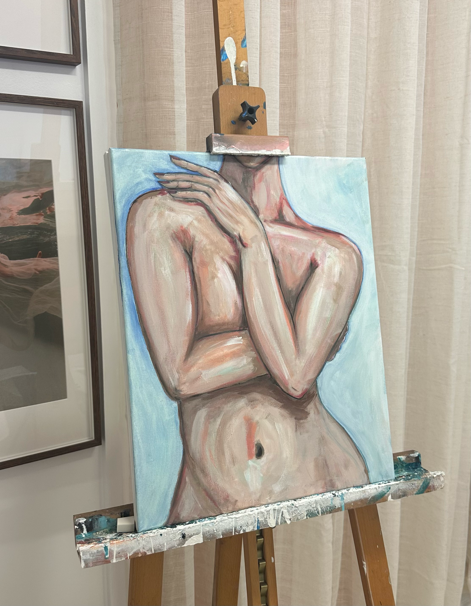 A nude figure painting in soft pastels sits on an easel against a light curtained background.