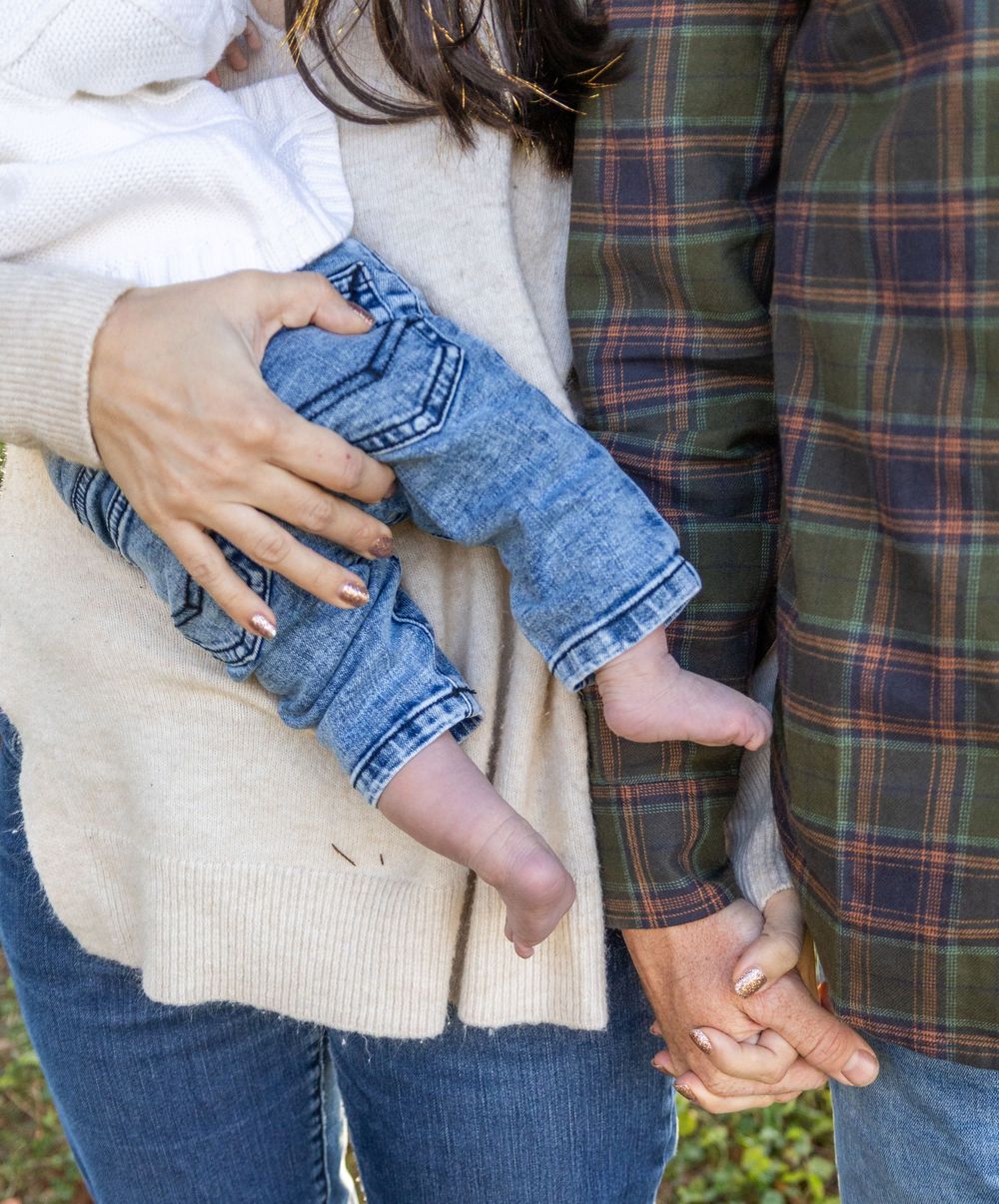 Close-up of parents holding newborn baby in jeans and white outfit while holding hands.