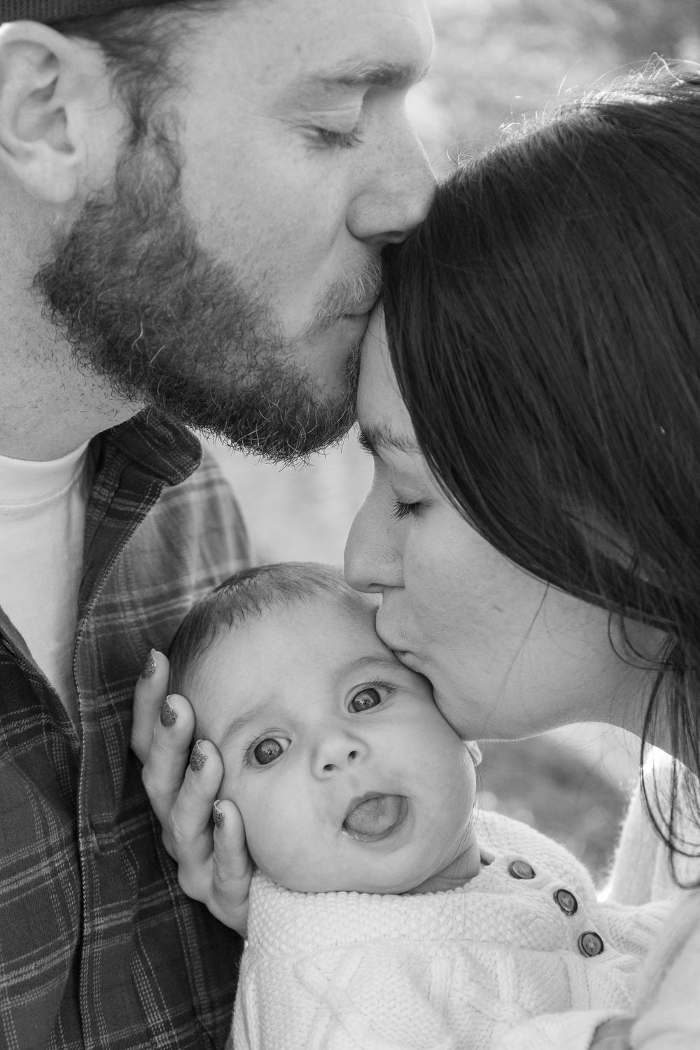 Black and white photo of parents kissing their wide-eyed newborn baby on both cheeks.