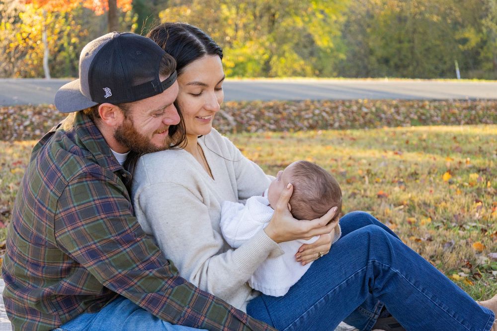 Young couple sitting outdoors in autumn sunlight lovingly cradling their sleeping newborn baby.