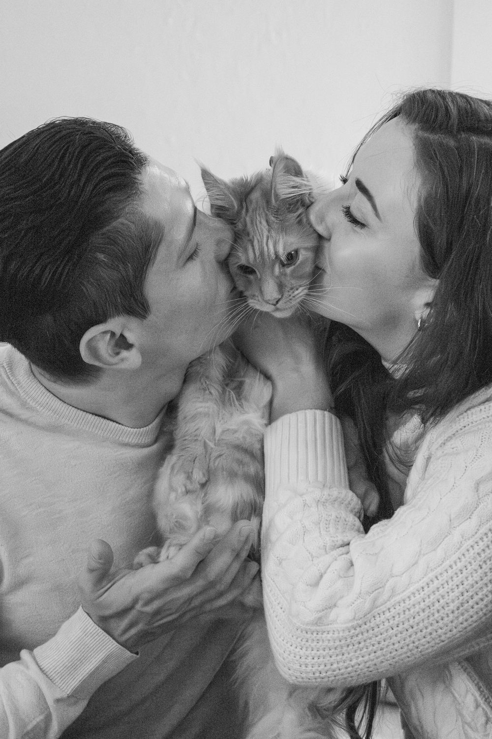 Black and white photo of couple kissing their fluffy ginger Maine Coon cat on both sides.