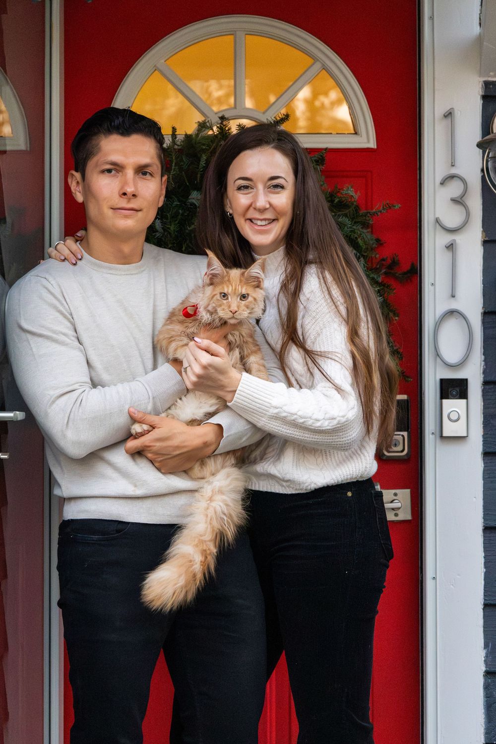 Smiling couple holding fluffy ginger Maine Coon cat in front of red Christmas decorated door.