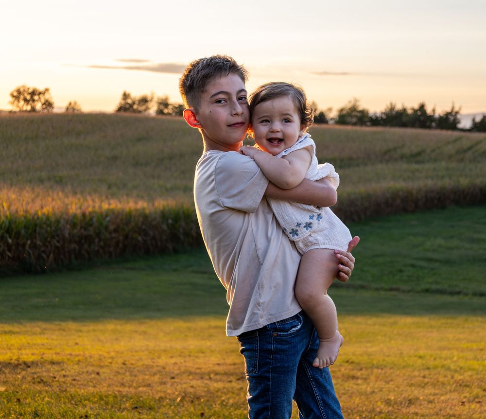 A boy hugs a smiling baby girl in a field at golden hour.