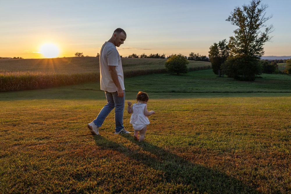 A father walks hand in hand with his toddler at sunset in a field.