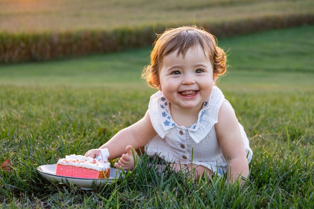 Smiling baby girl sitting on green grass with a slice of birthday cake on a plate during golden hour.
