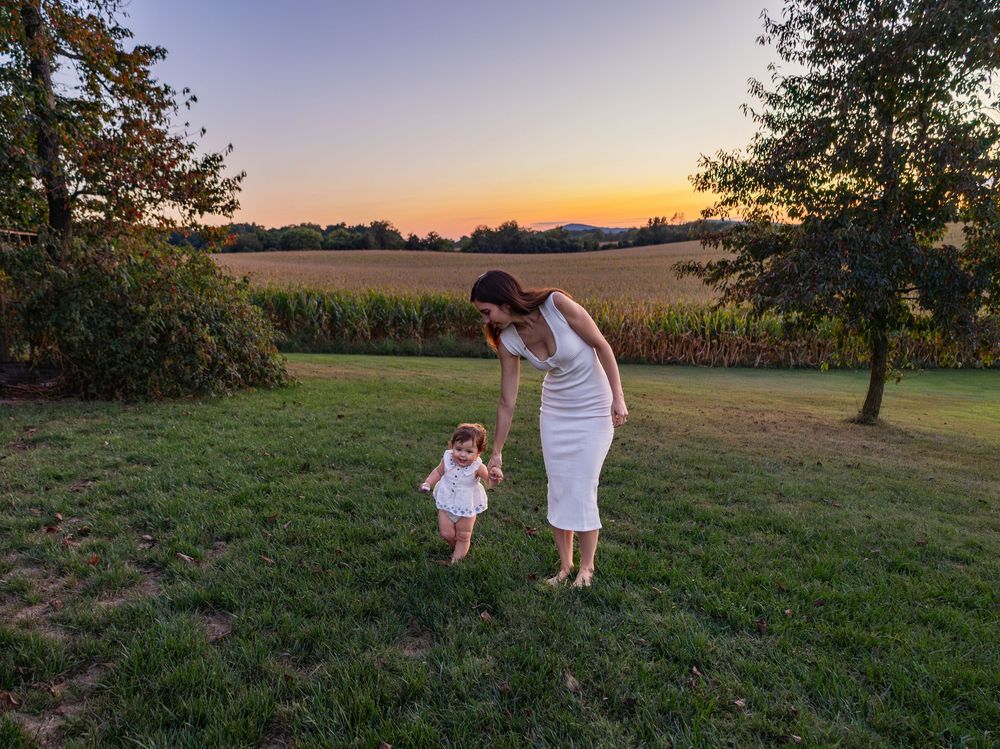Mother helping toddler walk on green grass at sunset with a cornfield in the background.