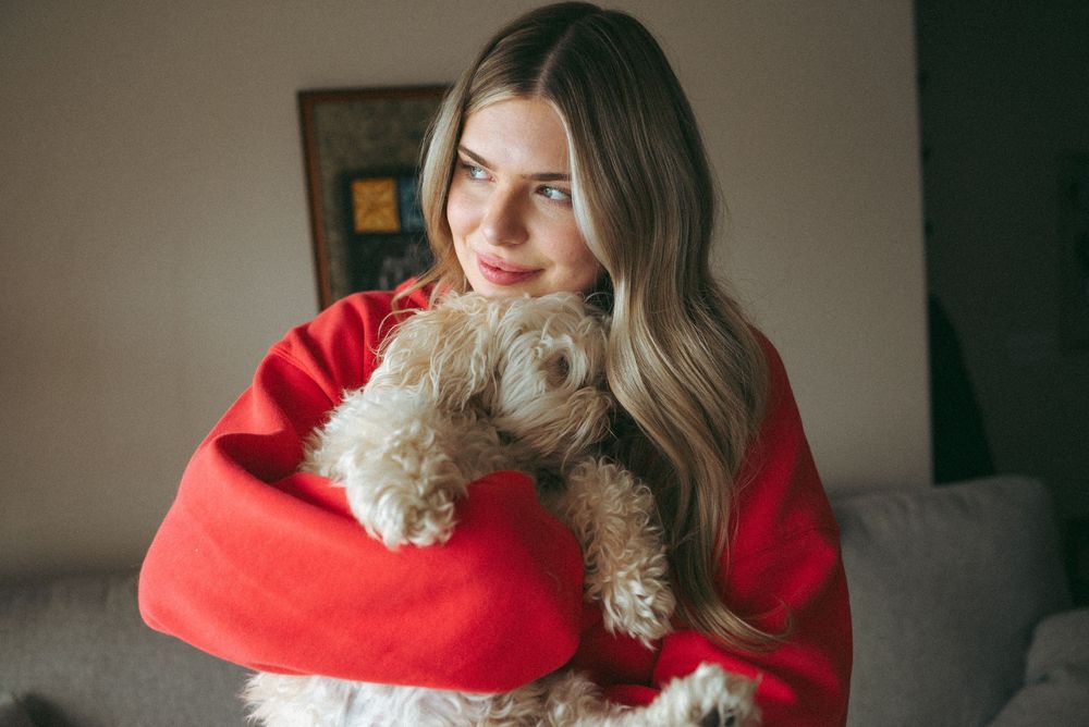 Someone wearing a bright red sweater cuddles with a small fluffy dog while sitting on a grey couch in soft lighting.