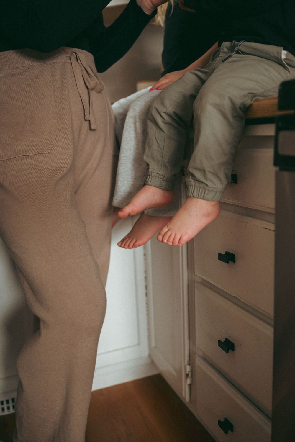 Bare feet dangling off white dresser drawers with casual beige pants in a cozy indoor setting.