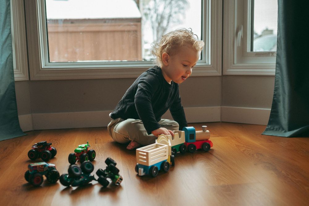 Child playing with wooden toy trains on hardwood floor by a sunny window.