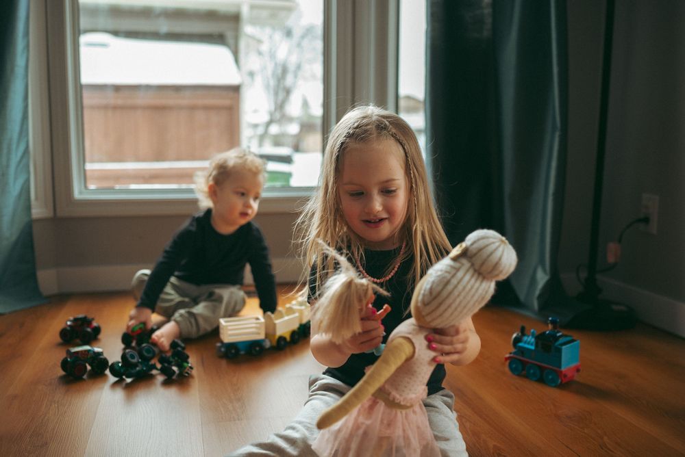 Two children play on a hardwood floor with toys, with natural light streaming through a window.