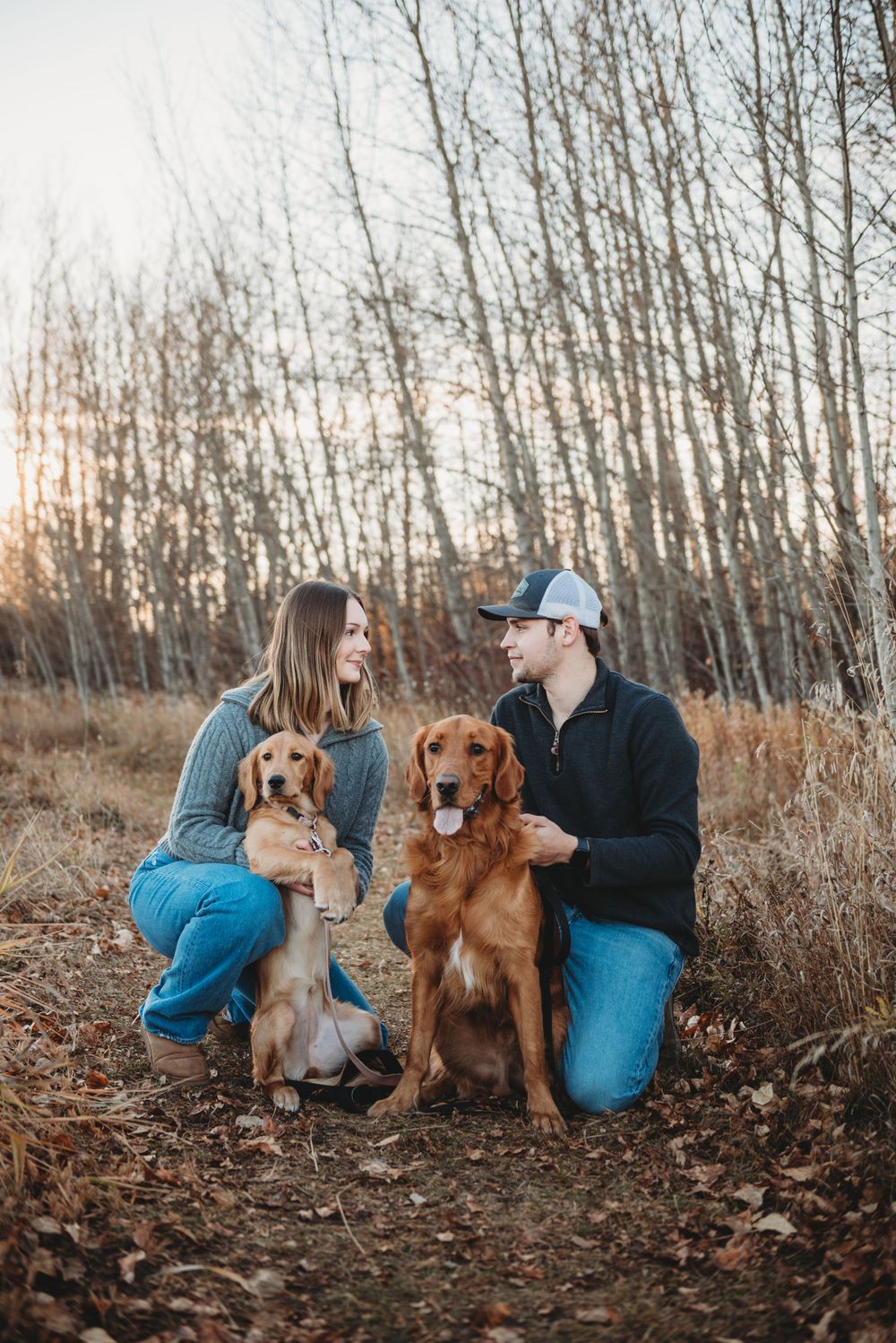 A couple sits in a forest with their two dogs - a golden retriever and another pup - during sunset amongst bare trees.