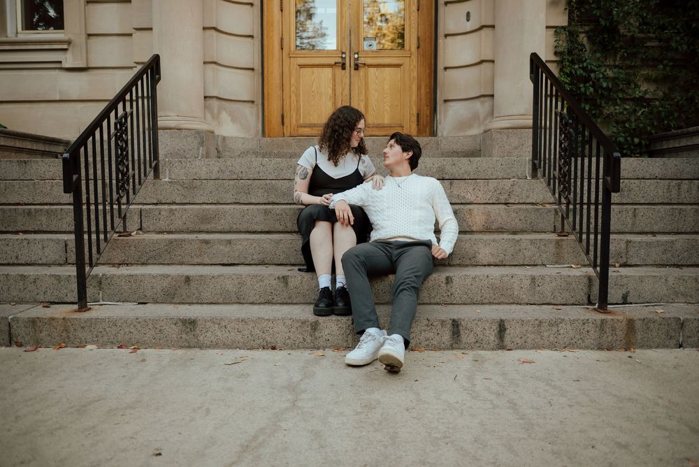 Two people sit together on concrete steps outside a stone building in a romantic moment.