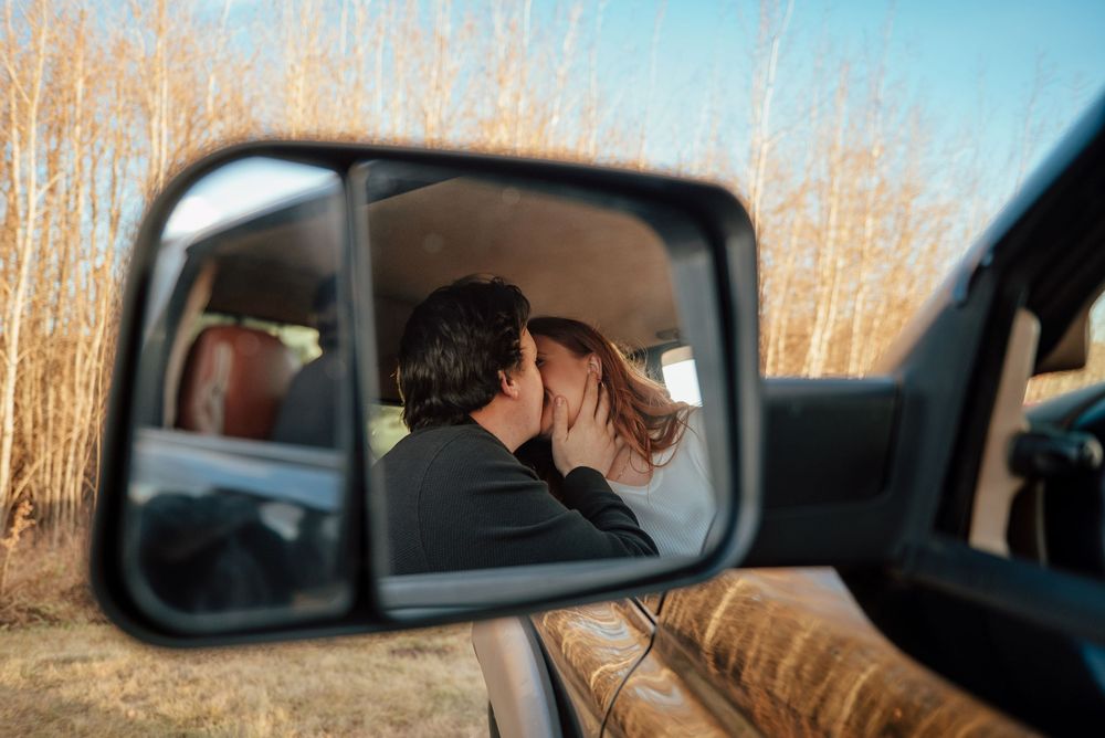 A couple sits together in a car while surrounded by bare winter trees reflected in the side mirror.