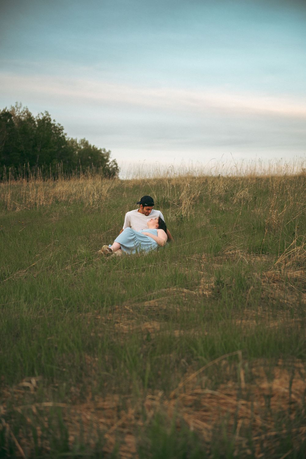 Someone in light clothing sits on grassy hill against moody sky at sunset in rural landscape.