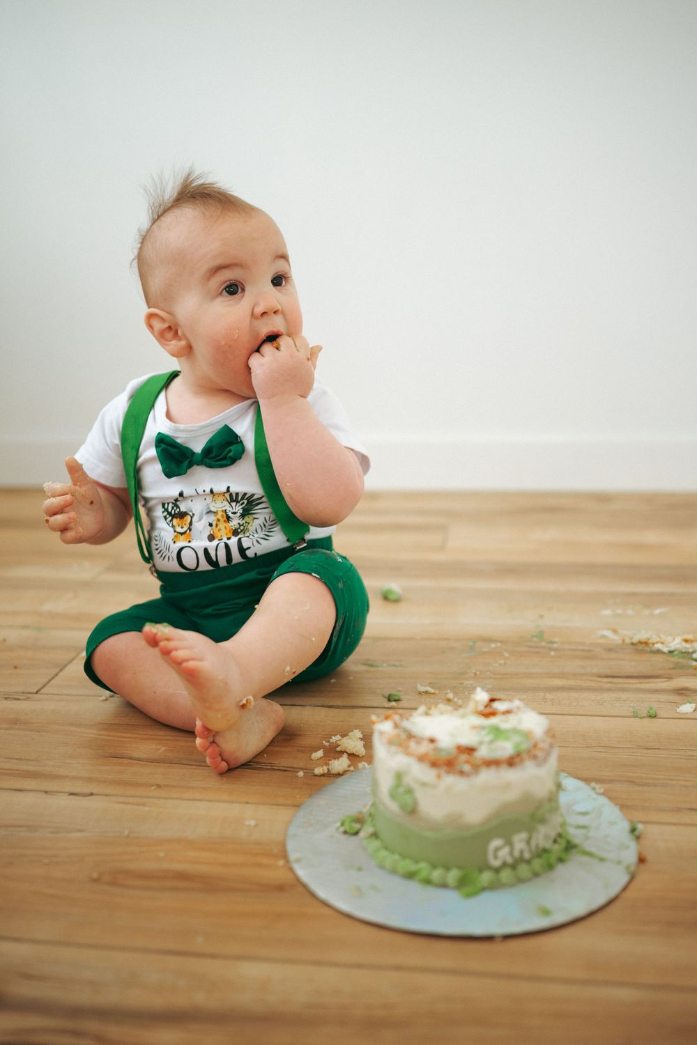 A baby in green overalls sits on hardwood floor next to a small smashed birthday cake during a cake smash photo session.