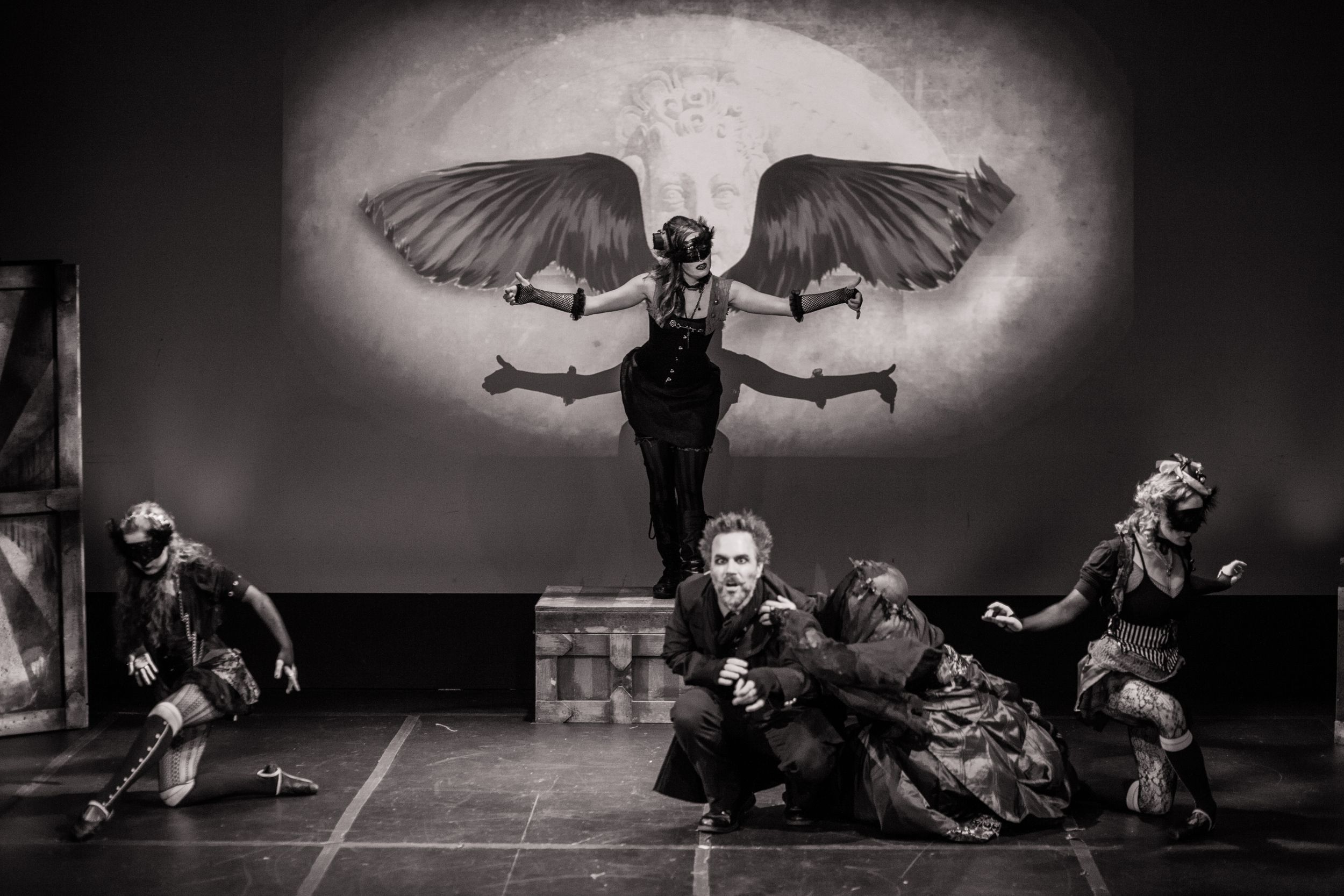 Theatrical performers in dramatic winged costume dance on stage in black and white.
