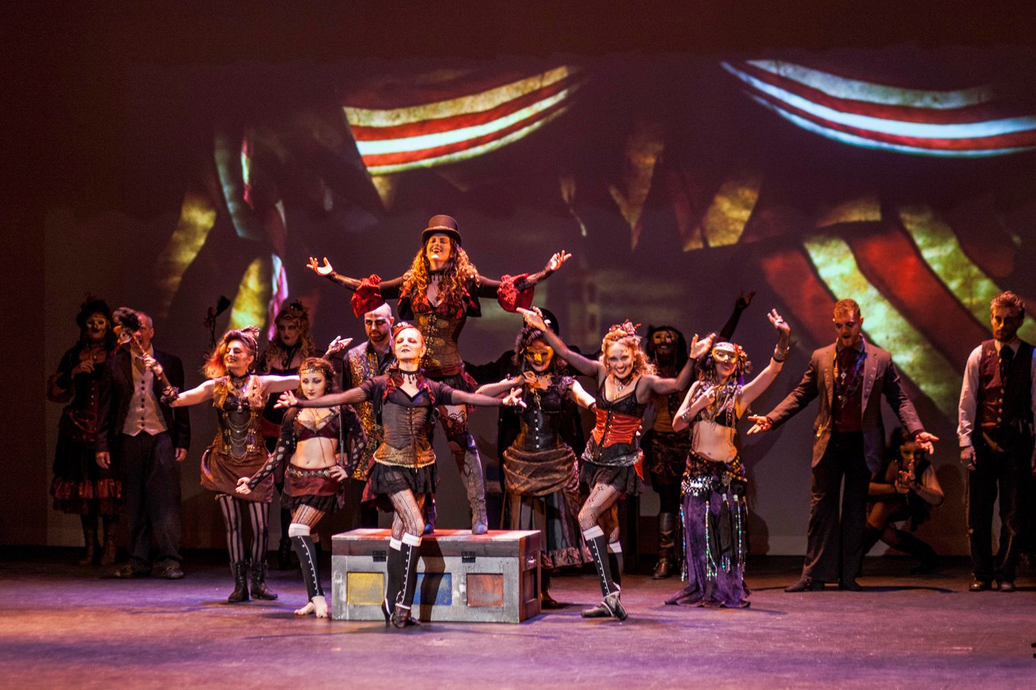 Theatrical performers in steampunk costumes performing an energetic dance number on stage with colorful lighting.