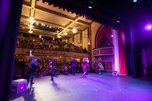 Wide view of a theater stage with performers under purple lighting and a full audience balcony.