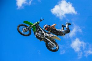 Freestyle motocross rider executes an aerial stunt with a green dirt bike against blue sky.
