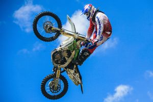 Dirt bike rider performs a vertical wheelie stunt against bright blue sky background.
