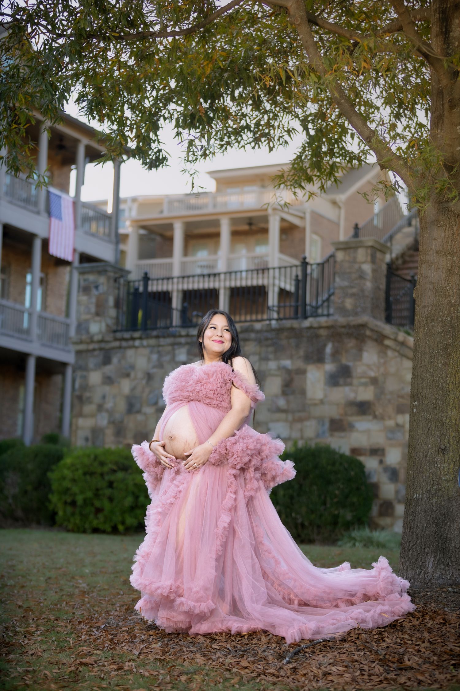 Maternity session in pink flowing gown against stone building and autumn trees.
