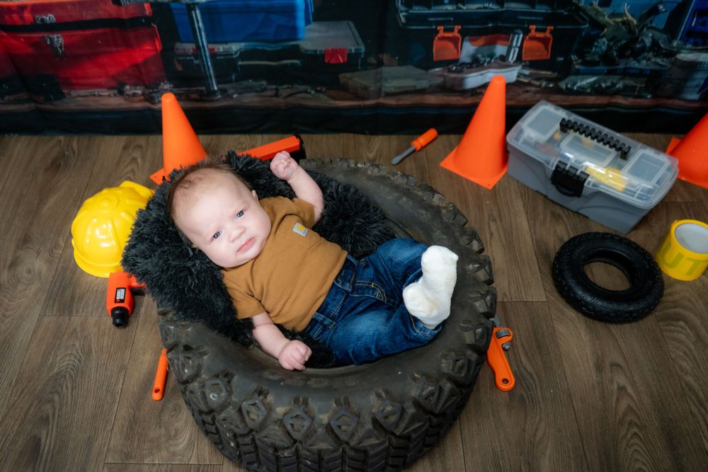 Small child rests comfortably inside a large tire surrounded by orange construction cones and tools on wooden floor.