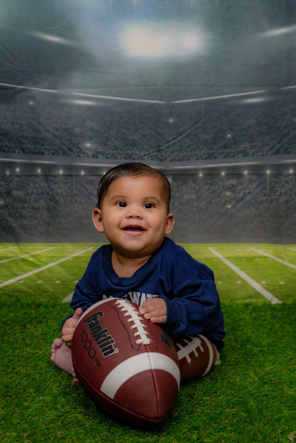 Young sports fan sits on football field turf with regulation football in studio portrait setting with stadium backdrop.