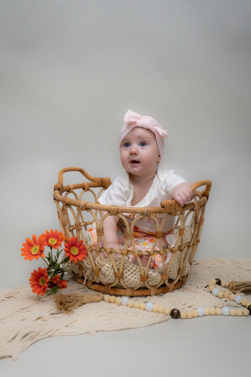 Baby in pink headband sits in woven basket with orange flowers and decorative beads on beige fabric backdrop.