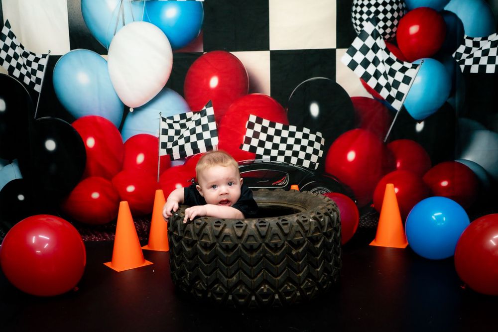 Race car themed party backdrop with checkered flags, red white and blue balloons, orange cones, and tire prop decorations.
