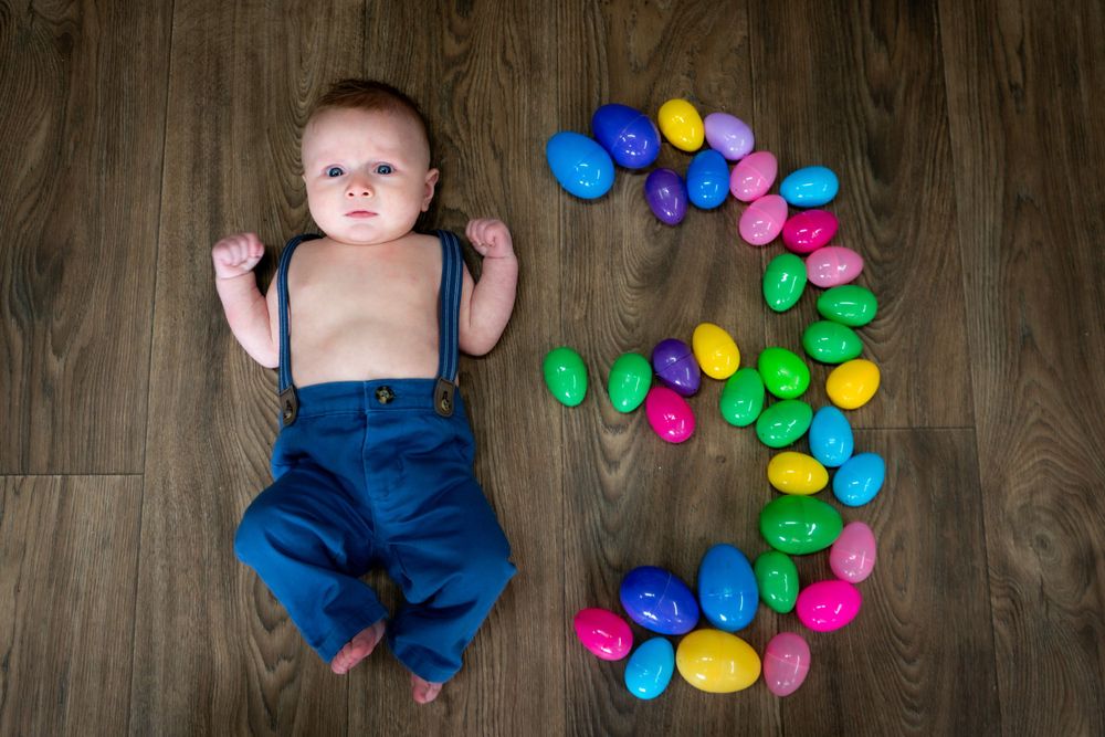 A baby laying on wooden floor surrounded by colorful plastic eggs arranged in the shape of number 3.