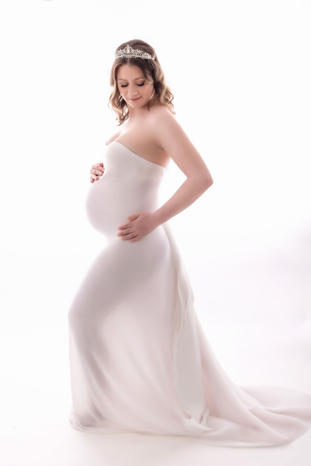 Elegant maternity photo featuring a white flowing gown and crystal tiara against a bright white background.