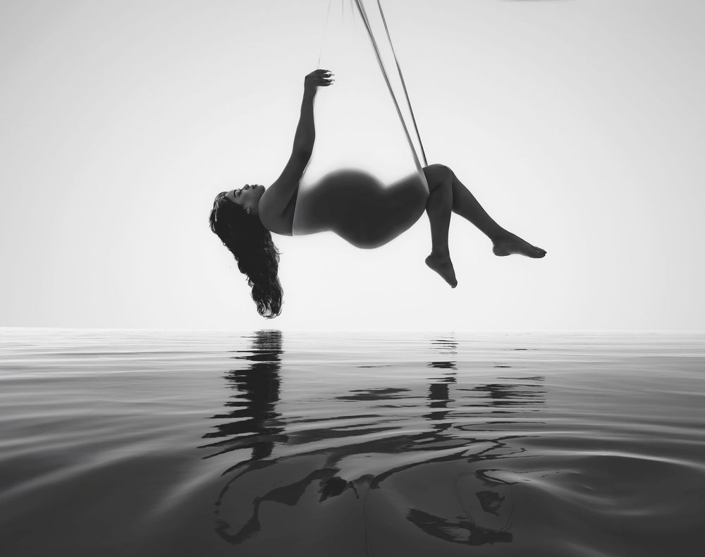 Black and white photo of a pregnant woman suspended on aerial silks above calm reflective water.