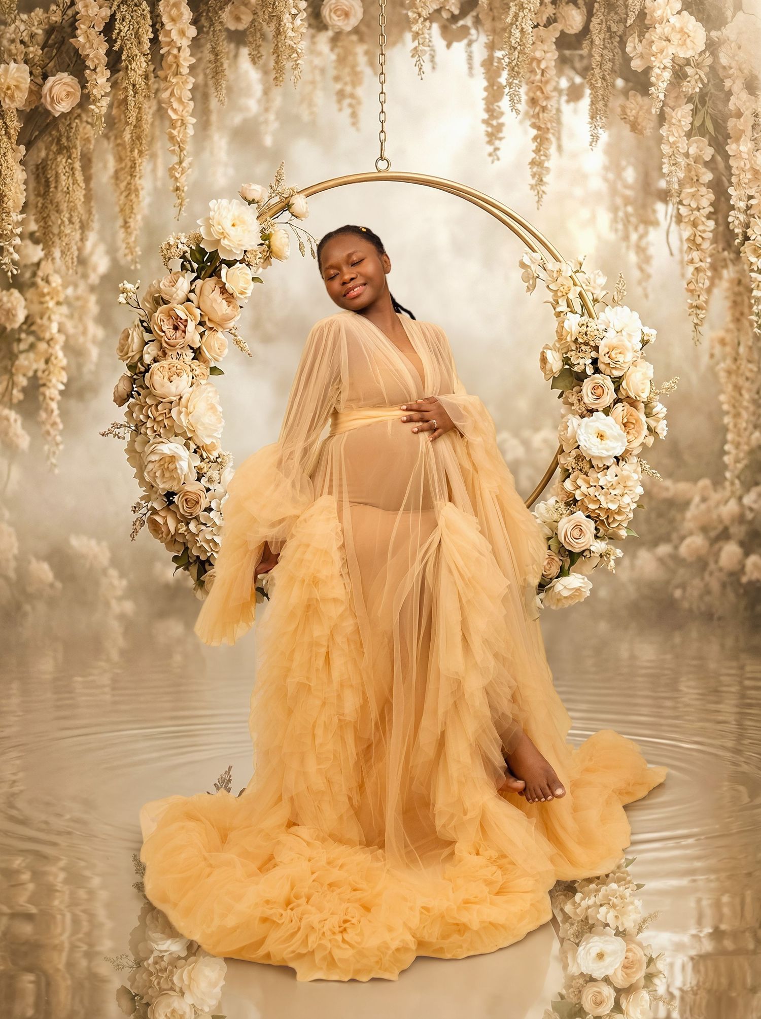 Pregnant woman in golden tulle gown poses on floral swing surrounded by white roses in dreamy maternity photo.