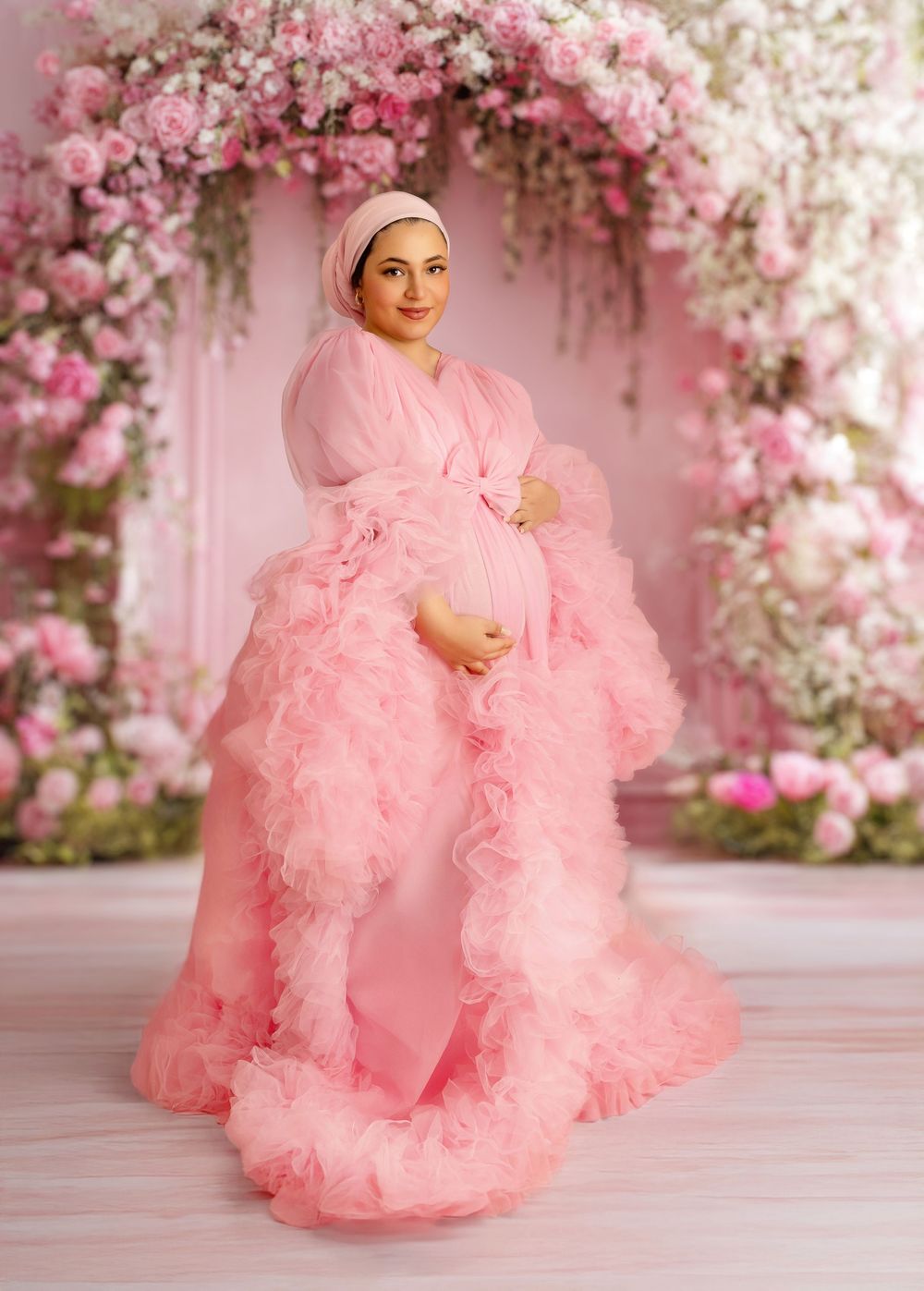 A dreamy pink maternity photoshoot with floral decorations and a flowing tulle gown in soft romantic lighting.