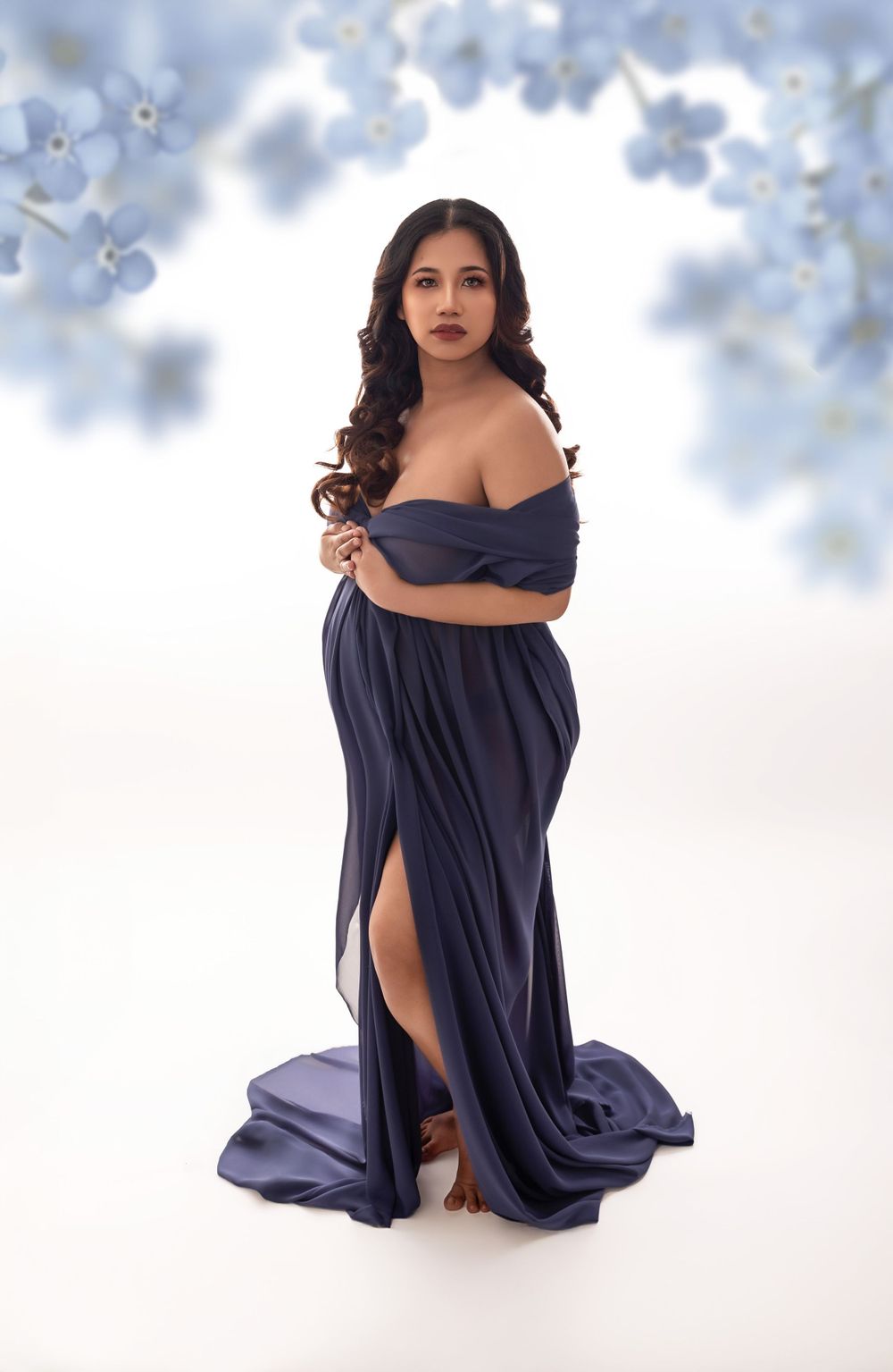 An elegant maternity photo featuring a navy blue off-shoulder flowing gown with a thigh-high slit against a soft cloudy backdrop.