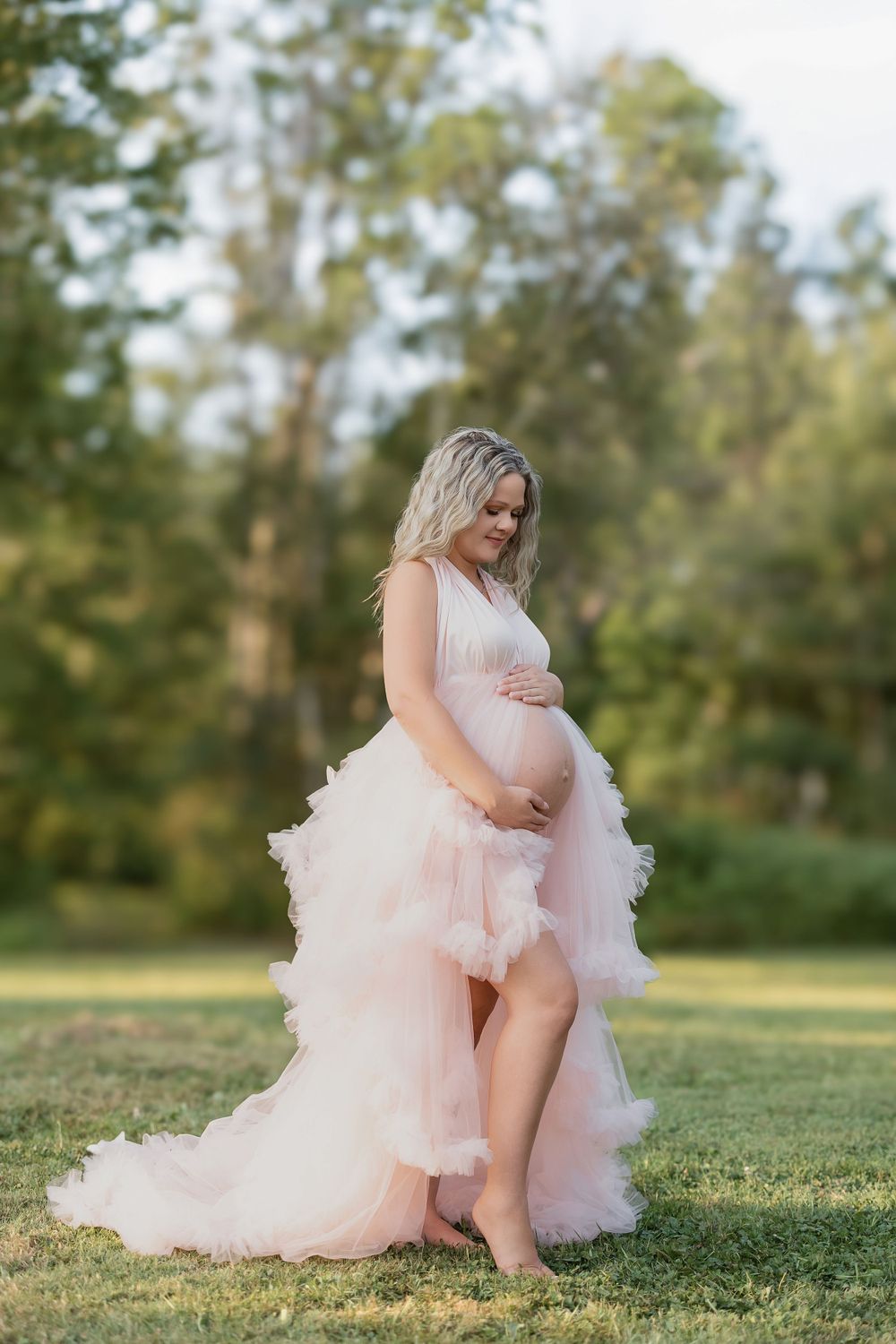 Ethereal maternity photo shows an expectant mother in a flowing blush tulle gown against a dreamy outdoor backdrop.