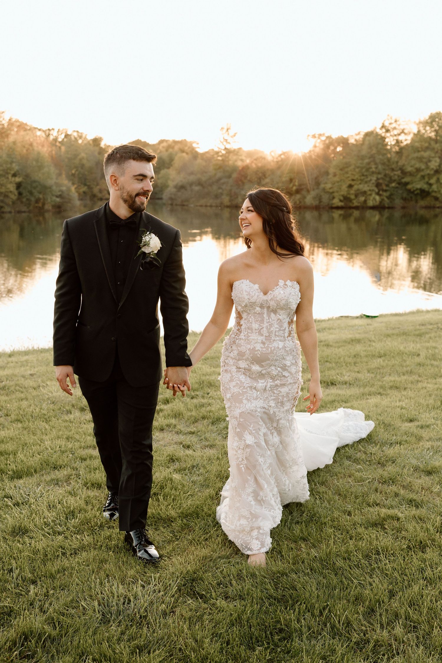 A couple walks hand in hand by a lake at sunset, with the bride wearing a lace wedding gown.