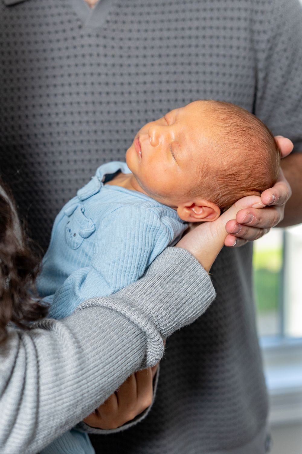 A sleeping newborn baby in a light blue outfit being cradled in by his parents in gray sweaters during a newborn photo session in their home