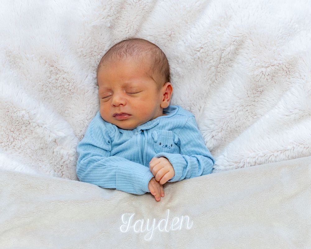 Sleeping newborn baby wearing soft blue sweater rests peacefully on white name blanket during a newborn photography session in Boston, MA.