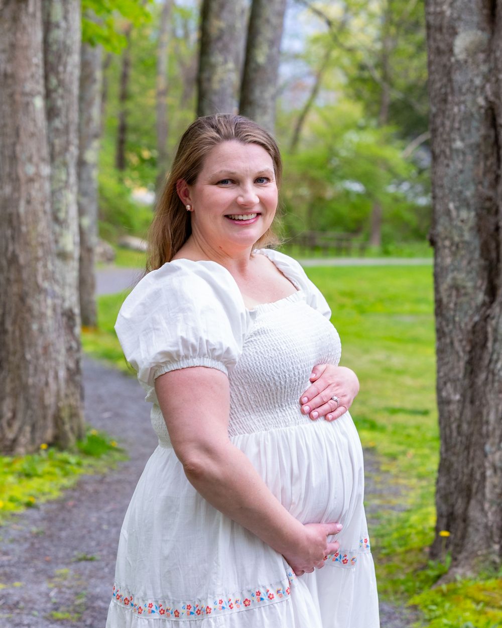 A person in a white dress with puff sleeves poses on a forest path during spring during a maternity photo portrait session outside Boston.