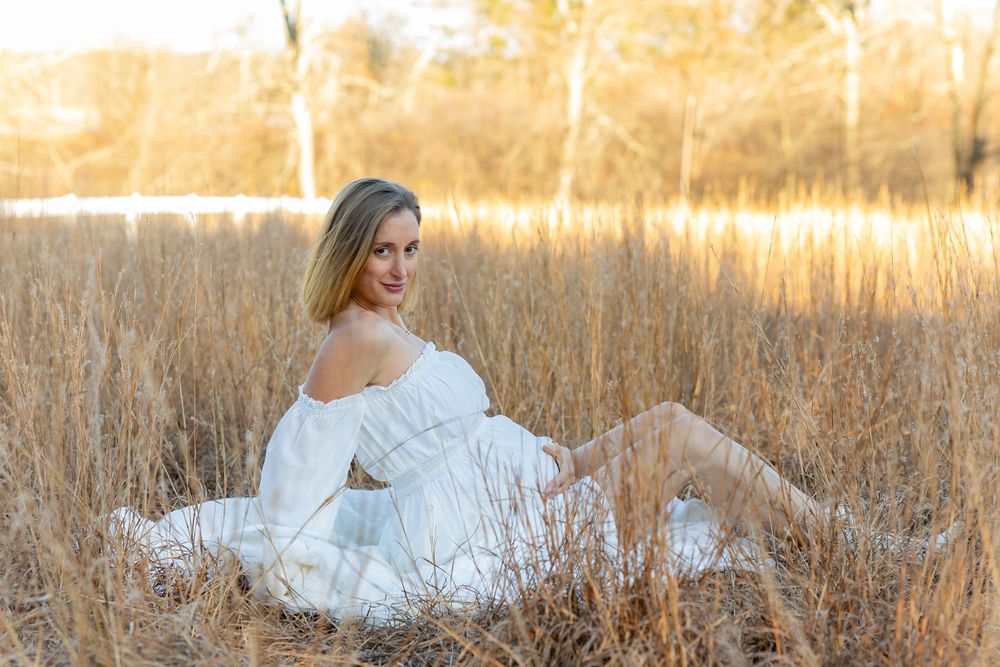 A figure in a flowing white off-shoulder dress poses gracefully in a golden field at sunset at a maternity portrait shoot in Massachusetts