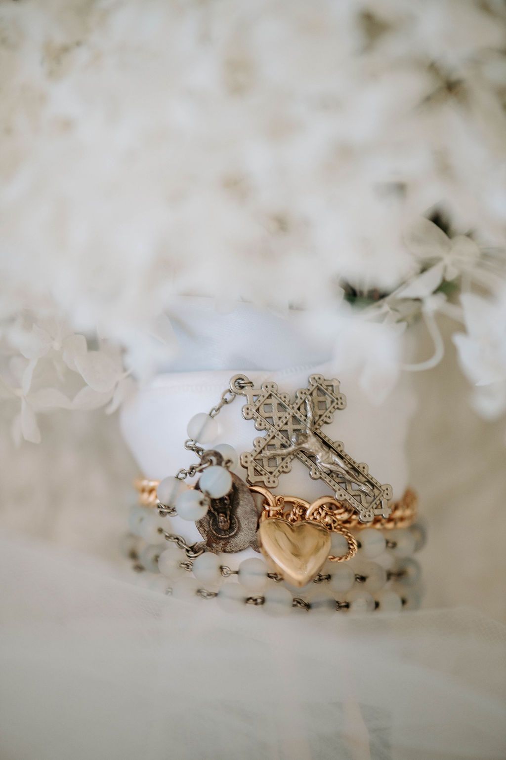 Delicate silver cross necklace and heart charm arranged on white surface with soft floral background.