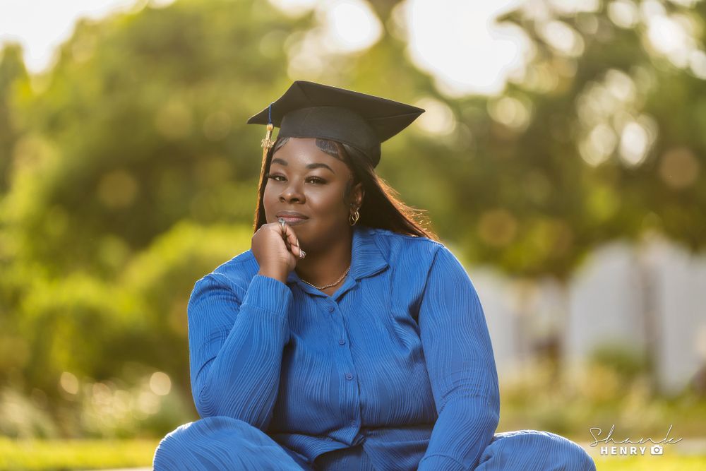 A graduate in blue attire sits thoughtfully on grass during a sunny graduation photoshoot.