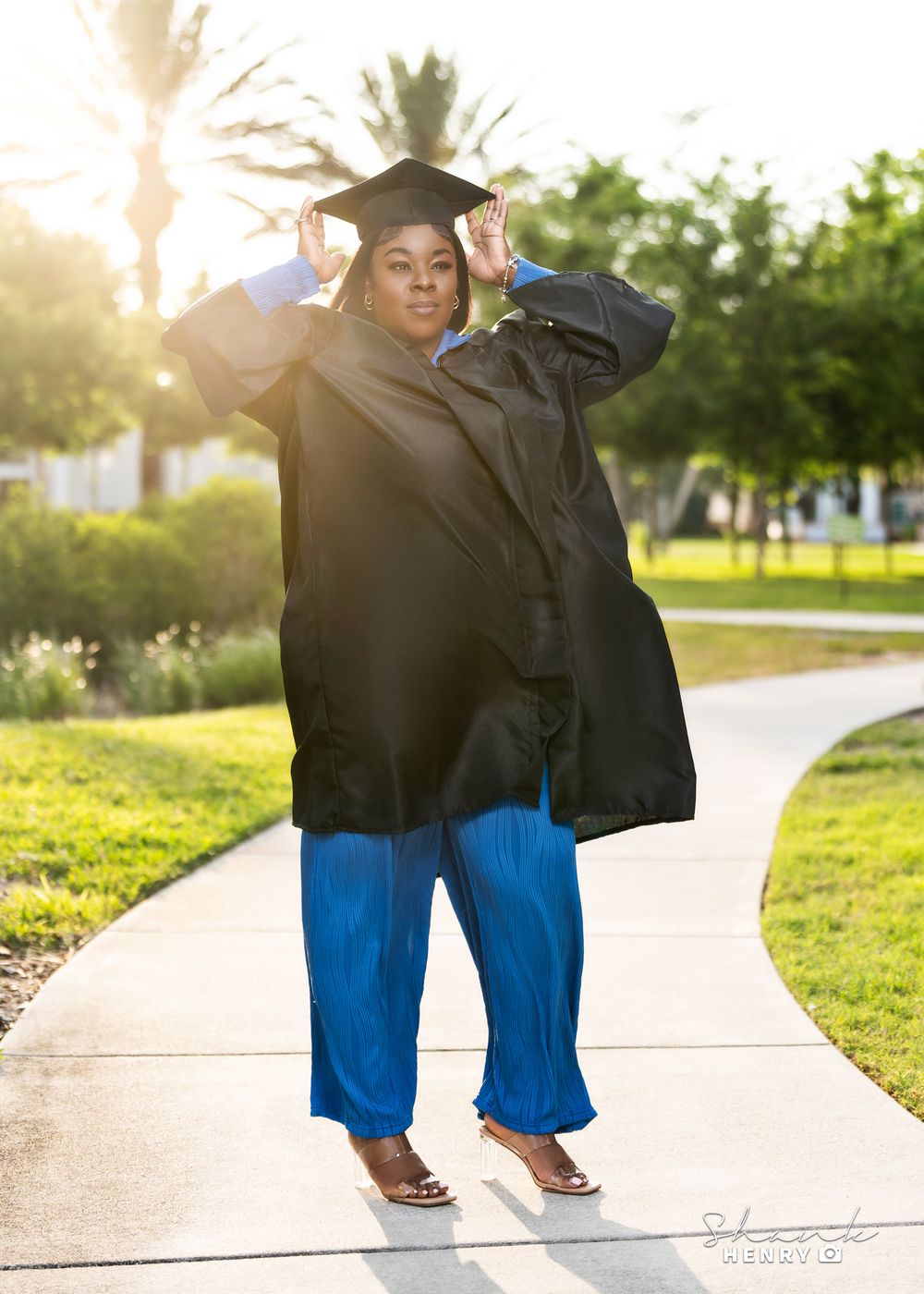A graduate in a black cap and gown strikes a joyful pose on a sunny pathway.
