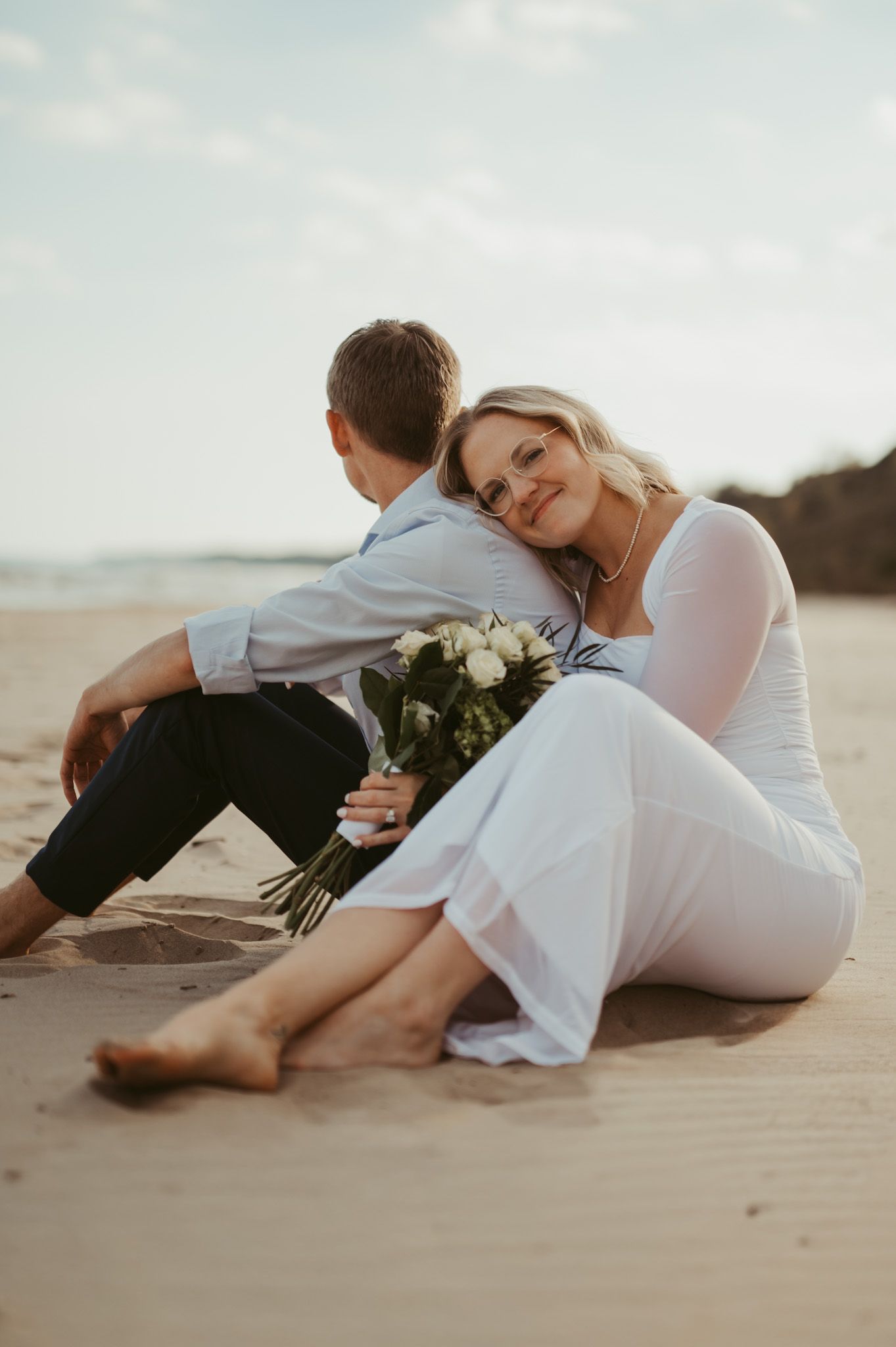 Couple sitting on sandy beach, bride in white dress leaning on groom, holding white flower bouquet, romantic lakeside elopement.