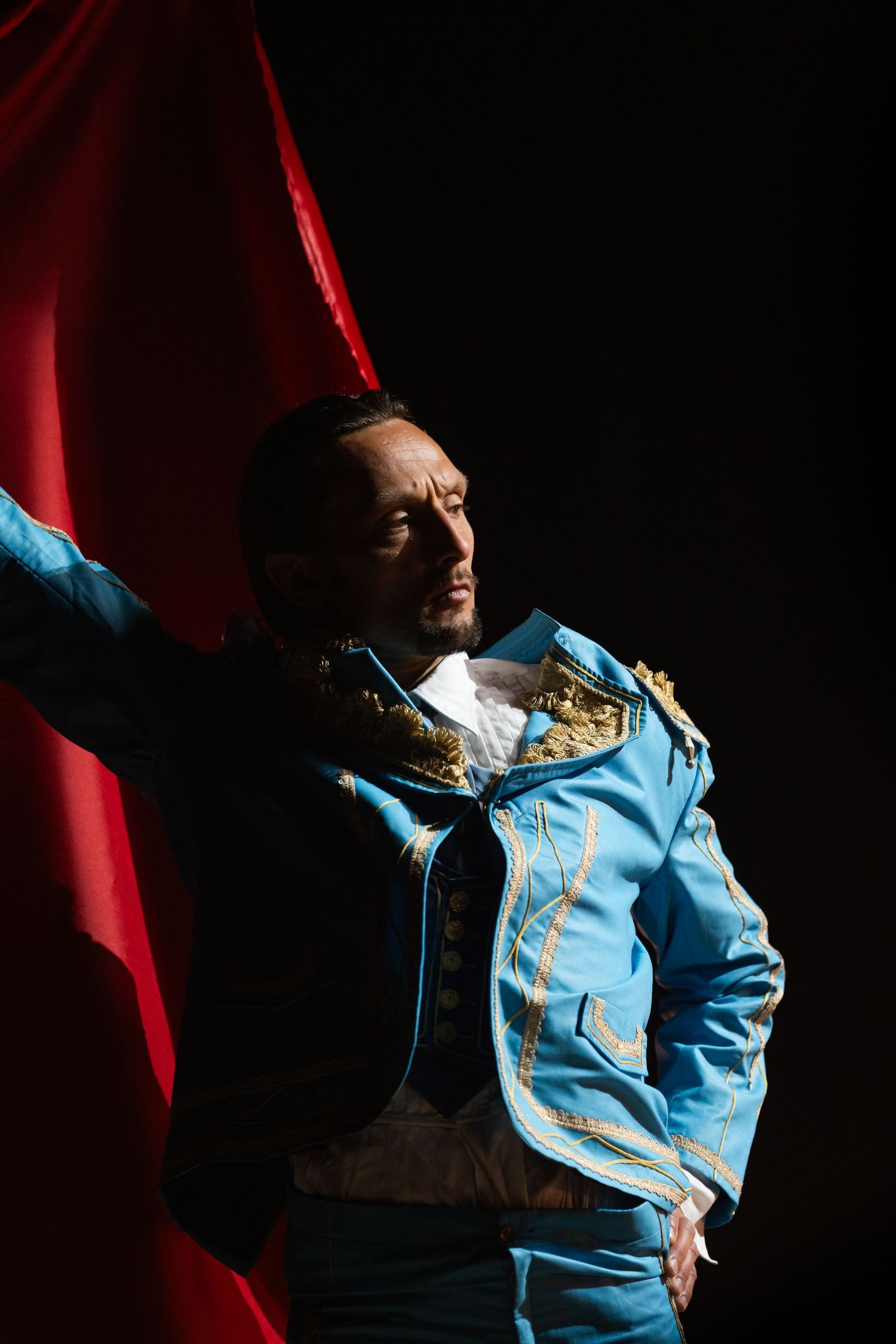 Performer in ornate blue and gold uniform holding red fabric in dramatic lighting.